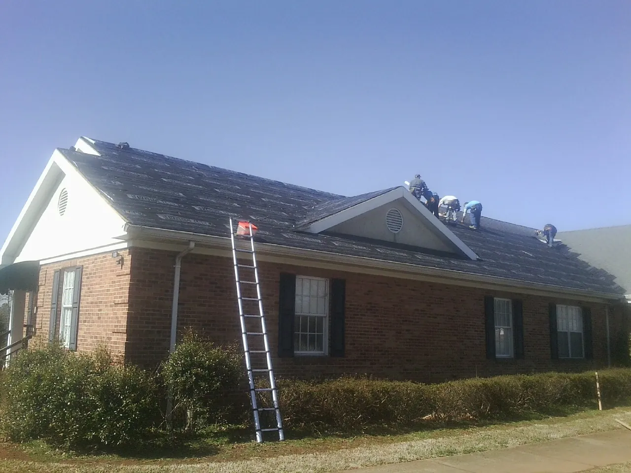 Crew tearing shingles off a brick ranch roof with a ladder set — Emergency Roof Repair work by our crew in Canastota