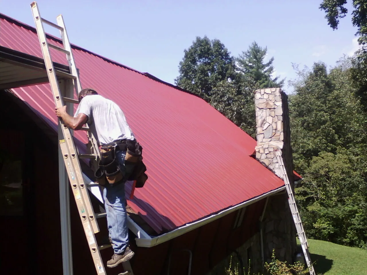 Roofer on a ladder beside a red metal roof and stone chimney — Soffit & Fascia Repair by Roofing Brothers in Canastota
